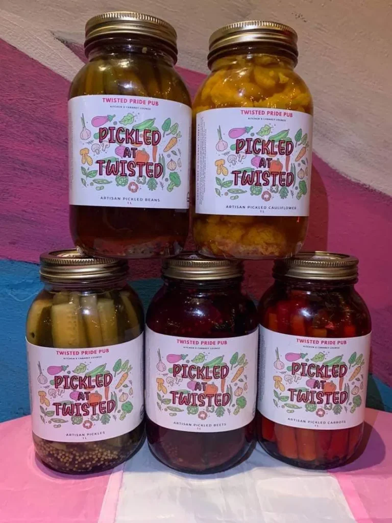 Five jars of colorful pickled vegetables are neatly stacked, each labeled attractively